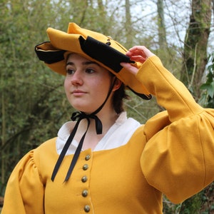 Puede incluir: Una mujer con un sombrero amarillo y negro, una chaqueta amarilla y una camisa blanca. El sombrero tiene un ala ancha y est&aacute; decorado con una cinta negra. La chaqueta tiene un cuello alto y mangas largas. La mujer est&aacute; de pie en un bosque.