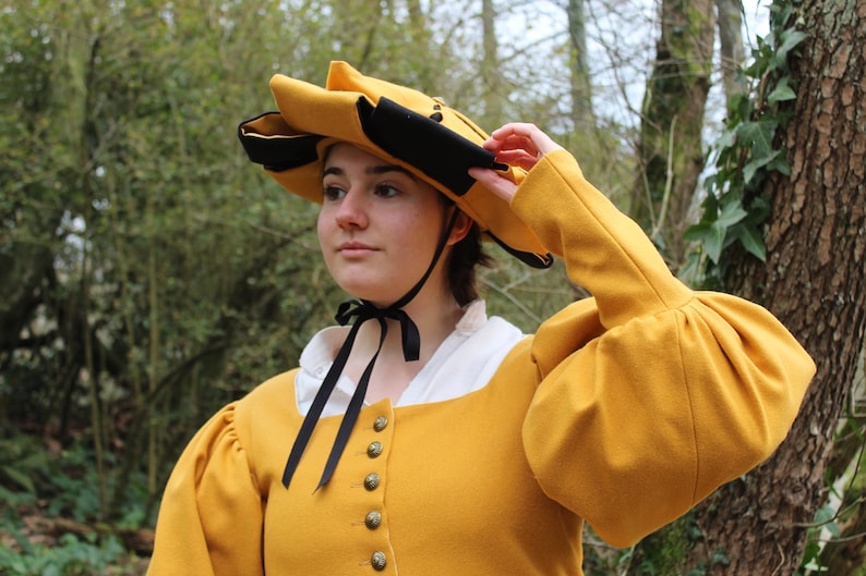 Puede incluir: Una mujer con un sombrero amarillo y negro, una chaqueta amarilla y una camisa blanca. El sombrero tiene un ala ancha y est&aacute; decorado con una cinta negra. La chaqueta tiene un cuello alto y mangas largas. La mujer est&aacute; de pie en un bosque.