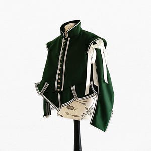 May include: A dark green jacket with white trim and accents, displayed on a mannequin. The jacket features a high collar, decorative buttons, and white ribbon details on the sleeves. The garment has a layered design with a ruffled hem.