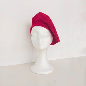 Puede incluir: Un gorro rojo de estilo beret sobre una cabeza de maniquí blanca. El gorro está hecho de un tejido suave y flexible.