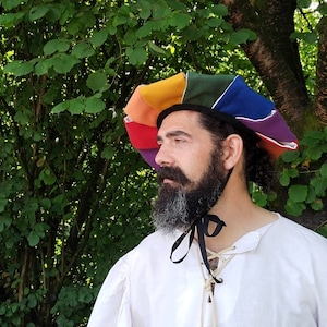 Puede incluir: Un hombre con un sombrero redondo multicolor y colorido con una cinta negra atada alrededor de la parte inferior. El sombrero está hecho de tela y tiene una variedad de colores, incluyendo rojo, naranja, amarillo, verde, azul y morado. El hombre tiene una barba larga y gris y lleva una camisa blanca.