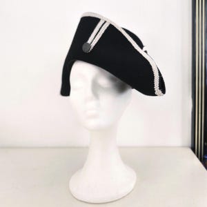 Könnte beinhalten: Ein schwarzer Zweispitzhut mit weißer Verzierung und einem dekorativen Knopf. Der Hut wird auf einem weißen Schaufensterkopf präsentiert. Das Design des Hutes weist eine gebogene Krempe und eine spitze Krone auf, typisch für historische Militär- oder formelle Kleidung.