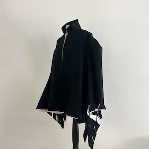May include: A black cape with a high collar and unique leaf-shaped cutouts at the hem. The cape is displayed on a black mannequin against a white background, showcasing its design and structure.