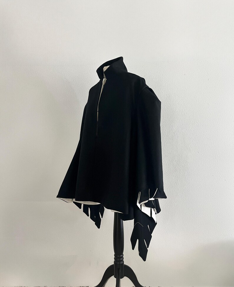 May include: A black cape with a high collar and unique leaf-shaped cutouts at the hem. The cape is displayed on a black mannequin against a white background, showcasing its design and structure.