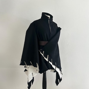 May include: A black cape-style garment with a high collar and a front zipper. The cape features a brown belt and white, jagged-edged accents along the hemline. The garment is displayed on a black mannequin against a white background.