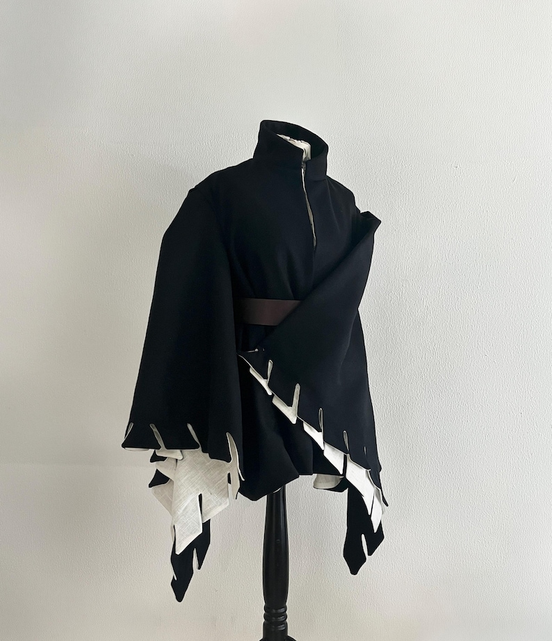 May include: A black cape-style garment with a high collar and a front zipper. The cape features a brown belt and white, jagged-edged accents along the hemline. The garment is displayed on a black mannequin against a white background.
