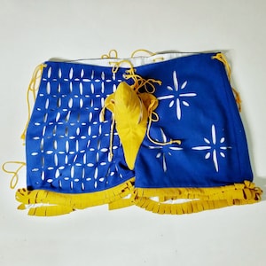 May include: Blue and white fabric shorts with yellow fringe trim. The shorts have a geometric pattern on one side and a star pattern on the other. A yellow fabric piece is attached to the center of the shorts.