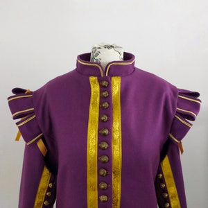 Puede incluir: Una chaqueta morada con detalles dorados, incluyendo ribetes y botones. La chaqueta presenta un cuello alto y hombros estructurados con detalles dorados. El ribete dorado recorre la parte delantera y alrededor del cuello. Esta prenda recuerda a la vestimenta histórica.