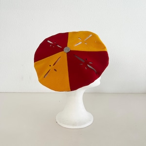 Op de afbeelding: Een ronde baret met afwisselende segmenten van donkerrode en goudgele stof. De muts is versierd met vier stervormige lasergesneden patronen en een centraal zilverkleurig rond knoopdetail boven op de kruin van de baret.