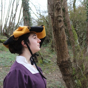 Puede incluir: Una mujer con un vestido morado y un sombrero negro y amarillo con una cinta negra atada al cuello. Est&aacute; de pie en un bosque.