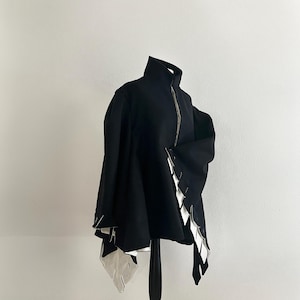 May include: A black jacket with a high collar and a silver zipper. The jacket features white, geometric accents along the edges, creating a striking contrast. The garment is displayed on a black mannequin, showcasing its unique design.