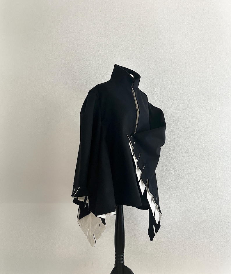 May include: A black jacket with a high collar and a silver zipper. The jacket features white, geometric accents along the edges, creating a striking contrast. The garment is displayed on a black mannequin, showcasing its unique design.