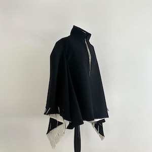 May include: A black cape with a high collar and a full-length zipper. The cape has a unique design with white and black geometric accents at the hem. The garment is displayed on a black mannequin against a white background.