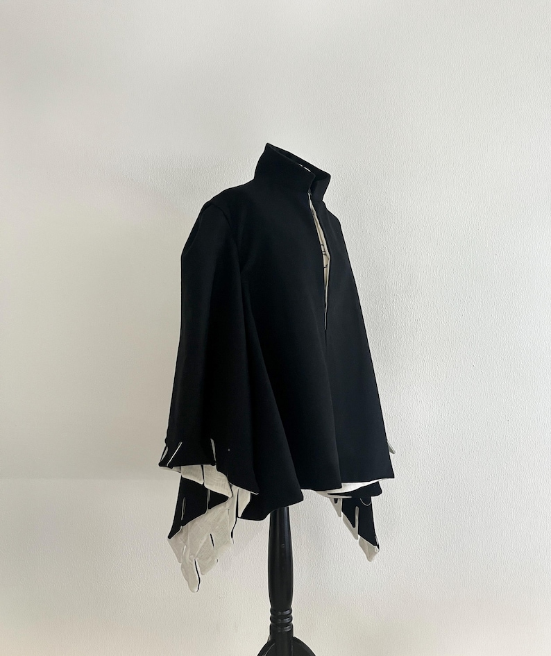 May include: A black cape with a high collar and a full-length zipper. The cape has a unique design with white and black geometric accents at the hem. The garment is displayed on a black mannequin against a white background.