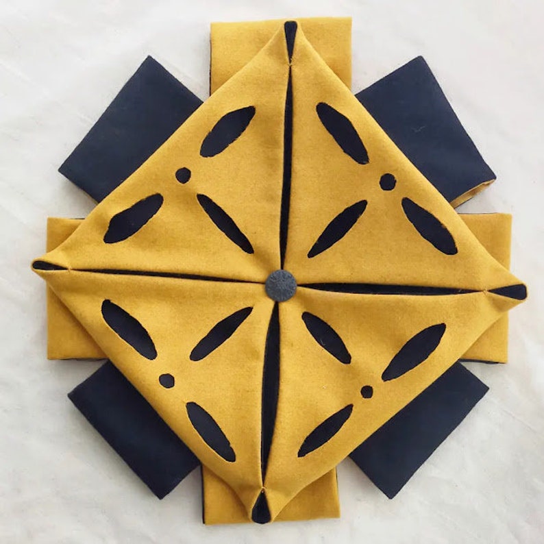 Puede incluir: Un cuadrado de tela amarillo con un dise&ntilde;o geom&eacute;trico negro, doblado en forma de estrella. La tela est&aacute; rodeada de cuadrados de tela negra.