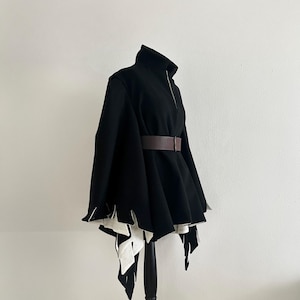 May include: A black cape-style jacket with a high collar and a brown belt. The jacket has a zipper closure and a unique design with white, jagged edges at the hem. The garment is displayed on a black mannequin.