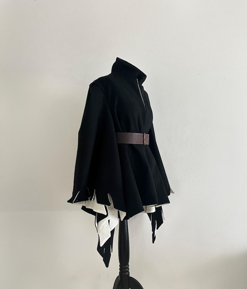 May include: A black cape-style jacket with a high collar and a brown belt. The jacket has a zipper closure and a unique design with white, jagged edges at the hem. The garment is displayed on a black mannequin.