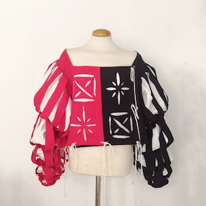 May include: A red and black cropped top with white geometric designs. The top has large, puffy sleeves with alternating red and white stripes.