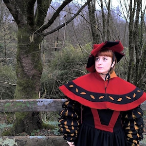 May include: A person wearing a red and black Renaissance-style costume with a matching hat. The costume features a red cape with black and gold accents, a black dress, and puffed sleeves with gold and black details. The background includes a wooden fence and trees.