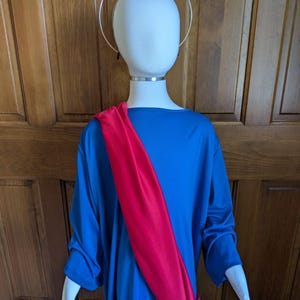 May include: A white mannequin head and torso wearing a blue tunic with a bright red sash draped diagonally across the chest. A thin, circular gold-toned halo is positioned above the head, secured by a band around the neck of the mannequin.