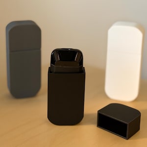 Könnte beinhalten: Ein schwarzes elektronisches Gerät mit abgerundeter Oberseite wird in einem schwarzen Gehäuse mit abgenommenem Deckel dargestellt. Zwei weitere Gehäuse, eines grau und eines weiß, sind ebenfalls vorhanden. Die Artikel befinden sich auf einer hellbraunen Oberfläche.
