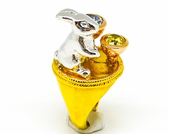 May include: A gold ring with a white rabbit figurine and two gemstones. The rabbit is holding a small cup with a yellow gemstone. The ring is on a white background.