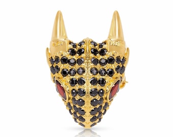 Puede incluir: Un broche dorado en forma de cabeza de dragón, con piedras de cristal negras y rojas. Los ojos del dragón son rojos y el resto del cuerpo está cubierto de piedras de cristal negras.