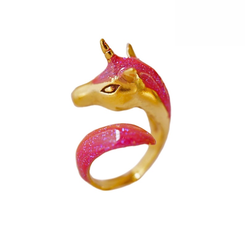 May include: A gold unicorn ring with pink glitter enamel. The unicorn's head is facing left and the horn is visible. The ring is made of metal and has a shiny finish.