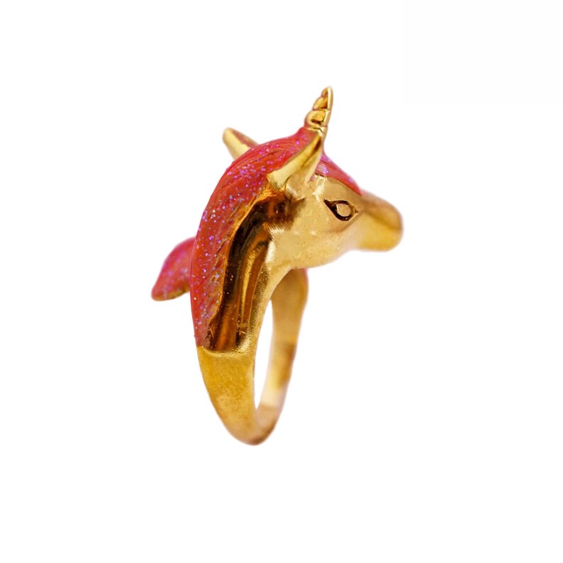 May include: Gold ring with a pink unicorn head design. The unicorn's horn is pointed upwards and the ring is made of metal.