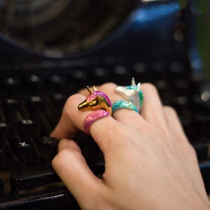 May include: Two unicorn rings, one pink and gold, the other blue and silver, worn on a person's finger.