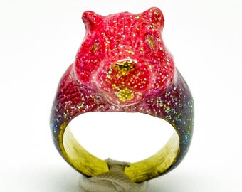 May include: A pink and purple glitter ring with a 3D animal head design. The ring is made of resin and has a gold band.