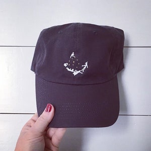 May include: A dark gray baseball cap with a white embroidered design featuring a person, stars, and a landscape. The cap has a curved brim and a fabric strap closure. The hat is held by a person.