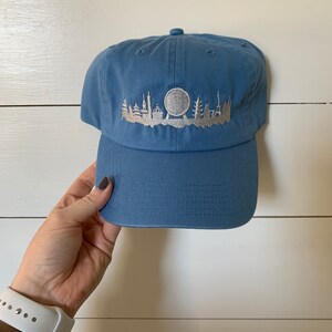 K&ouml;nnte beinhalten: Eine blaue Baseballkappe mit einer gestickten wei&szlig;en Silhouette einer Stadtlandschaft mit einem Riesenrad.