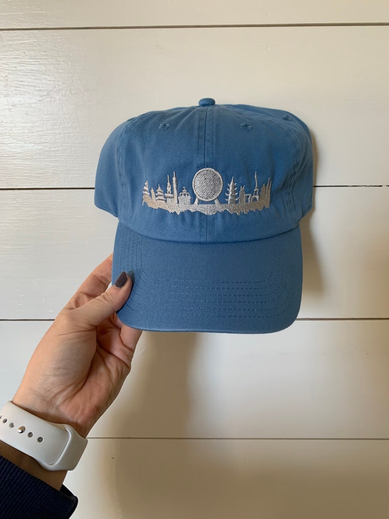 K&ouml;nnte beinhalten: Eine blaue Baseballkappe mit einer gestickten wei&szlig;en Silhouette einer Stadtlandschaft mit einem Riesenrad.
