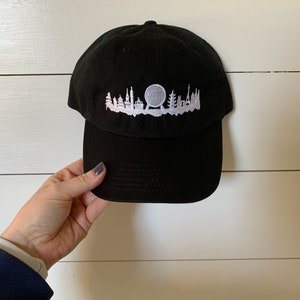 K&ouml;nnte beinhalten: Schwarze Baseballkappe mit einem wei&szlig;en gestickten Design einer Stadtlandschaft mit einem Riesenrad.