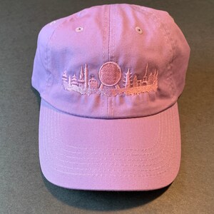 K&ouml;nnte beinhalten: Eine Baseballkappe in Lavendelfarbe mit einem gestickten Skyline- und einem Riesenrad-Design.