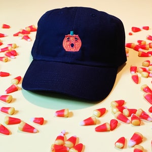 May include: A black baseball cap featuring an embroidered orange pumpkin with a green stem and a smiling face. The cap is surrounded by candy corn.