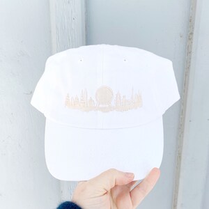 K&ouml;nnte beinhalten: Wei&szlig;e Baseballkappe mit einem beigefarbenen gestickten Stadtbild einer Stadt mit einer gro&szlig;en Kugel in der Mitte.