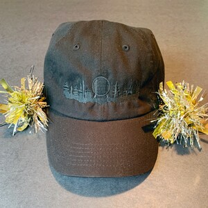 K&ouml;nnte beinhalten: Eine schwarze Baseballkappe mit einem gestickten Design einer Skyline und einer Kugel. Die Kappe hat einen gebogenen Schirm und einen schwarzen verstellbaren Riemen.