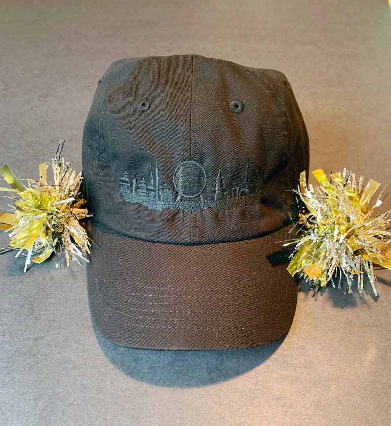 K&ouml;nnte beinhalten: Eine schwarze Baseballkappe mit einem gestickten Design einer Skyline und einer Kugel. Die Kappe hat einen gebogenen Schirm und einen schwarzen verstellbaren Riemen.