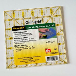 Könnte beinhalten: Ein transparenter, quadratischer Omnigrid-Lineal, 15,24 cm x 15,24 cm, mit gelben Gitterlinien und Messungen. Das Lineal enthält Winkel zum Schneiden und Zuschneiden von Stoff. Text auf dem Etikett weist auf die Herstellung in den USA hin.