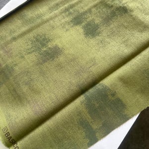 Puede incluir: Un trozo de tela verde doblado con un sutil patrón de camuflaje. La tela tiene una superficie texturizada y una etiqueta que dice "BASIC GREY / GREY".