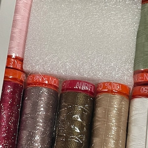 Pode incluir: Um conjunto de 10 carretéis de fio em várias cores, incluindo rosa, vermelho, cinza, verde, bege e branco. Os carretéis são envoltos em plástico transparente e têm etiquetas laranja com o texto "Aurifil 50%  Cott  " impresso neles.
