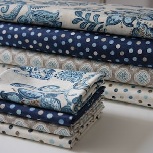 Könnte beinhalten: Ein Stapel Stoffquadrate in verschiedenen blauen und beigefarbenen Mustern, darunter Paisley, Punkte und geometrische Muster. Der Stoff ist ordentlich gefaltet und auf einer weißen Oberfläche angeordnet.