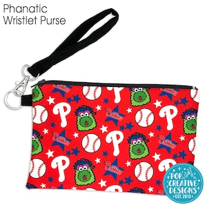 May include: Red wristlet purse with a black wrist strap. The purse has a zipper closure and features a repeating pattern of baseballs, stars, the letter "P", and a green furry creature. The text "Phillies" appears on the purse. The text "POP CREATIVE DESIGNS +EST. 2013+" is printed on the bottom right corner of the purse.