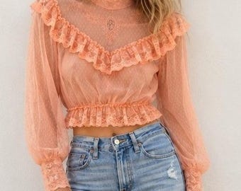 Op de afbeelding: Een perzikkleurige blouse met lange mouwen van doorschijnende kanten stof met een hoge kraag, ruches over de borst en schouders en een geplooide zoom. De blouse wordt gedragen met een lichtblauwe spijkerbroek.