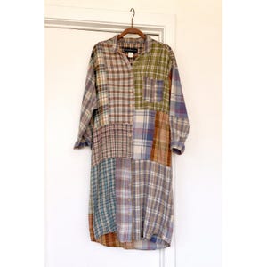 Könnte beinhalten: Ein langärmliges Patchwork-Hemdkleid in verschiedenen Karomustern. Das Kleid hat einen Kragen, eine Knopfleiste vorne und eine Mischung aus Farben wie Blau, Grün, Braun und Beige. Das Kleid hängt an einem Holzkleiderbügel.