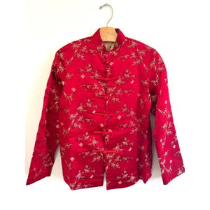 May include: A red, long-sleeved jacket with a mandarin collar and frog closures. The jacket features a gold bamboo and floral pattern. The garment is hanging on a wooden hanger against a white background.
