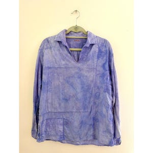 May include: A long-sleeve shirt in a light purple and blue tie-dye pattern. The shirt features a collared neckline with a V-shaped opening, a single patch pocket on the lower left side, and a second patch pocket on the upper right chest area.