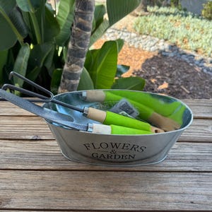 May include: An oval galvanized metal planter with the text FLOWERS & GARDEN printed on the front. Inside the container are two gardening tools with bright green handles and light wood accents, featuring metal heads with a textured finish.